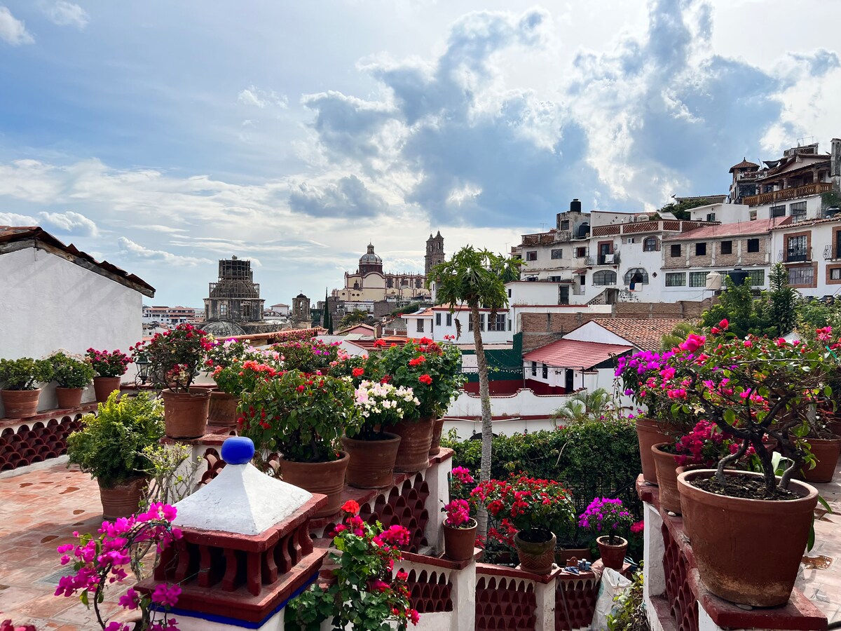 A panoramic view from a terrace is showcased, featuring vibrant flower pots filled with bougainvillea in shades of pink and purple. The background reveals charming buildings of Taxco, highlighted by the iconic structures of La Parroquia de Santa Prisca and San Sebastián under a partly cloudy sky.