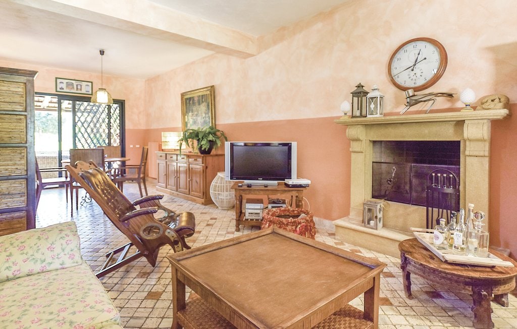 A comfortable living area is furnished with a wooden rocking chair and a coffee table. A television is positioned near the fireplace, which adds a warm touch. Light streams through a large window, and soft peach-colored walls enhance the inviting atmosphere.