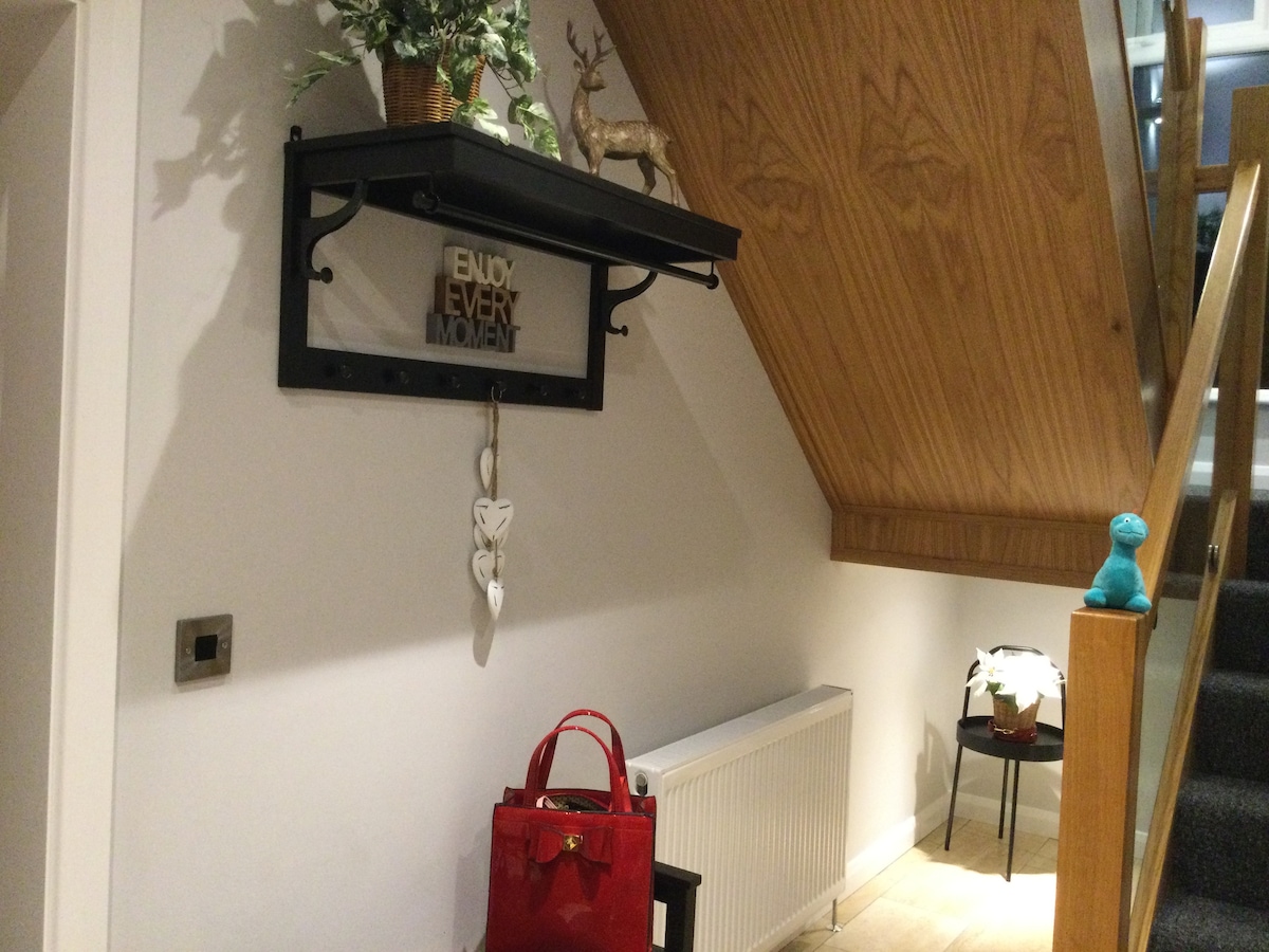 A well-organized entrance features a wall-mounted shelf displaying a decorative sign and plant. A stylish red handbag is placed on the floor beside a small table. The staircase with wooden accents leads to the upper living areas, complemented by soft lighting.