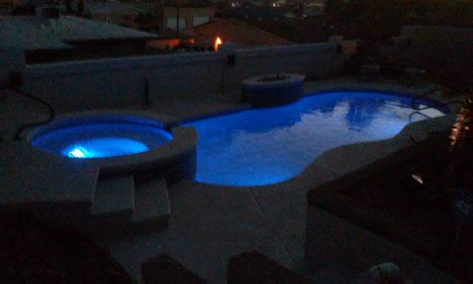 An illuminated pool area is visible at dusk, featuring a spacious swimming pool and a heated spa. Soft blue lighting reflects off the water's surface, creating a serene ambience. The surrounding space offers easy access to seating and outdoor features near the pool.