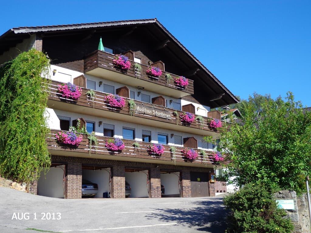 A multi-level building features several balconies adorned with vibrant flower boxes. Greenery surrounds the entrance, and two vehicles are parked beneath the main structure. Clear blue skies provide a bright backdrop to the scene, enhancing the building's inviting appearance.
