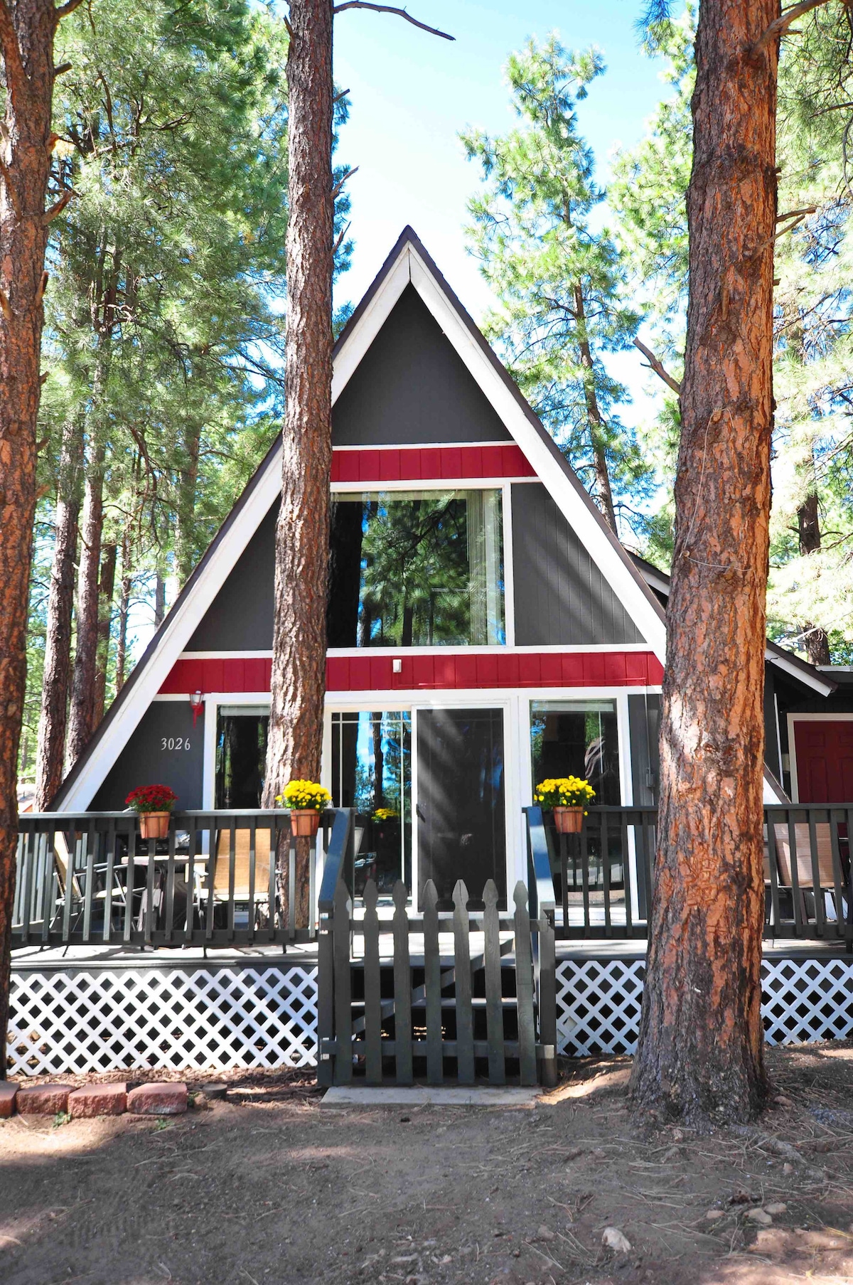 An A-frame cabin stands tall among ponderosa pines, featuring a striking black and red exterior. Large windows offer glimpses of the inviting interior. The front porch is adorned with flowers and surrounded by a white picket fence, creating a welcoming entrance.