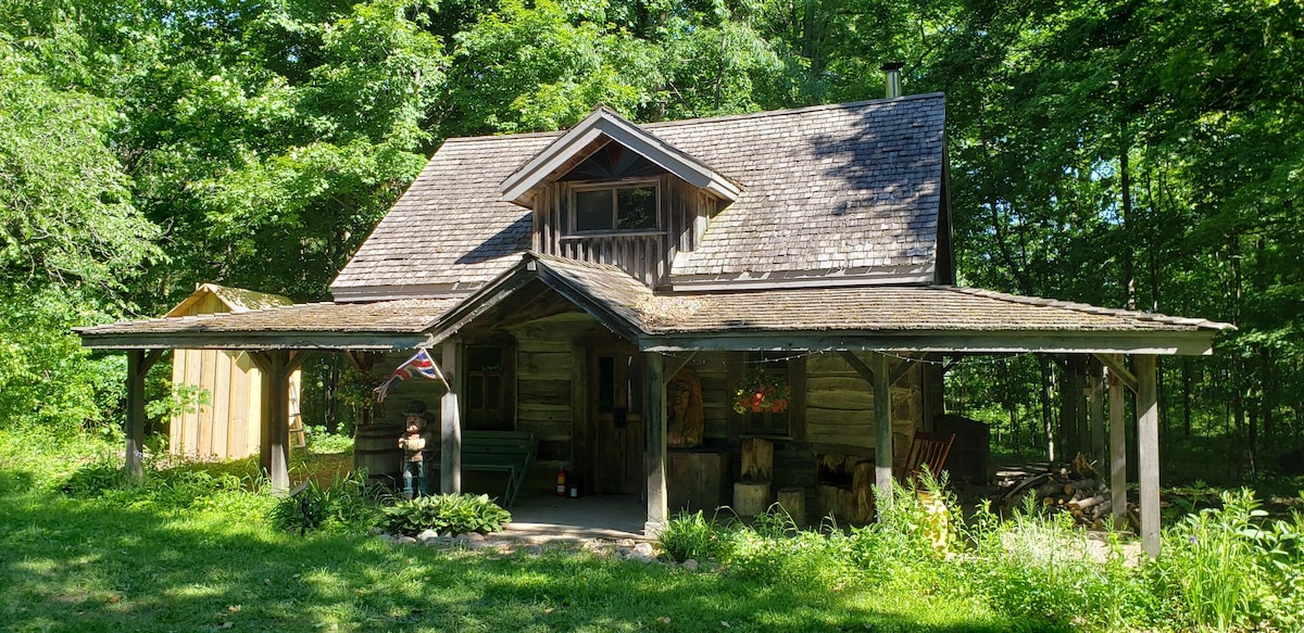 The log cabin is surrounded by lush greenery, showcasing a rustic wooden structure with a sloped roof. A covered porch features a rocking chair and a door leading inside, while a small storage area is visible to the left. The natural setting provides a serene, secluded atmosphere.