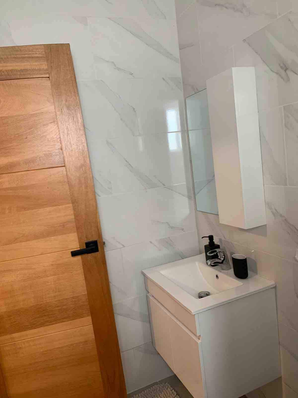 A modern bathroom features light-colored marble walls with a wooden door. A sleek white vanity with a sink is positioned against the wall, accompanied by a wall-mounted mirror above it. A dark container and a soap dispenser are placed on the countertop.