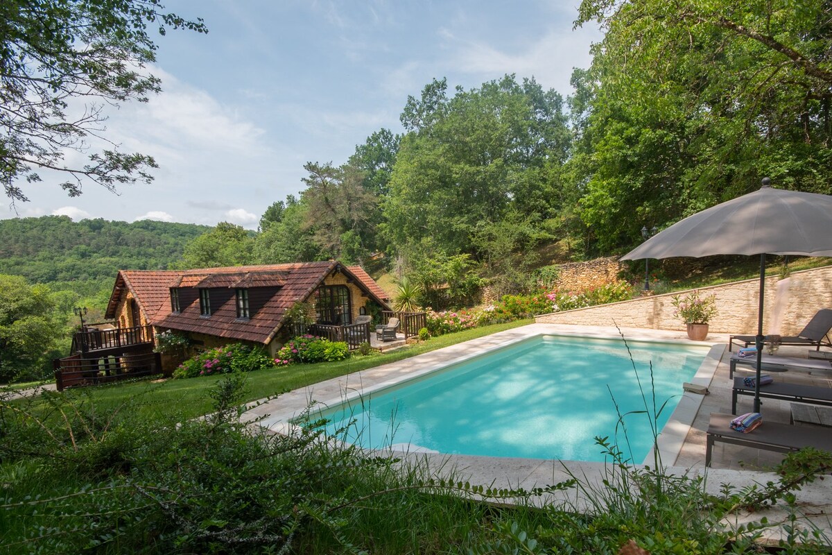 A serene outdoor setting features a clear swimming pool surrounded by lush green landscaping. A charming wooden house is visible in the background, along with comfortable lounge chairs and a sun umbrella near the pool, creating an inviting space for relaxation.