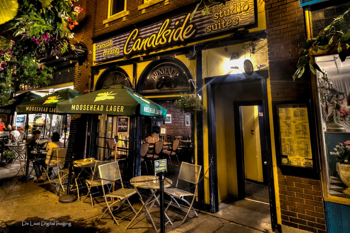A charming entrance is showcased with outdoor seating consisting of several metal chairs and tables, surrounded by vibrant greenery. The building features a colorful sign indicating 'Canalside Studio Suites,' complemented by decorative lighting and welcoming atmosphere.