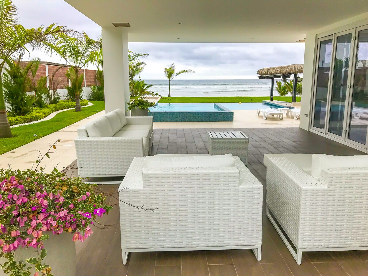 A spacious outdoor seating area is arranged with white woven furniture, facing a tranquil pool and the ocean beyond. Lush greenery surrounds the area, complemented by pink flowers in a nearby planter. The inviting space offers a relaxing atmosphere for enjoying the coastal views.