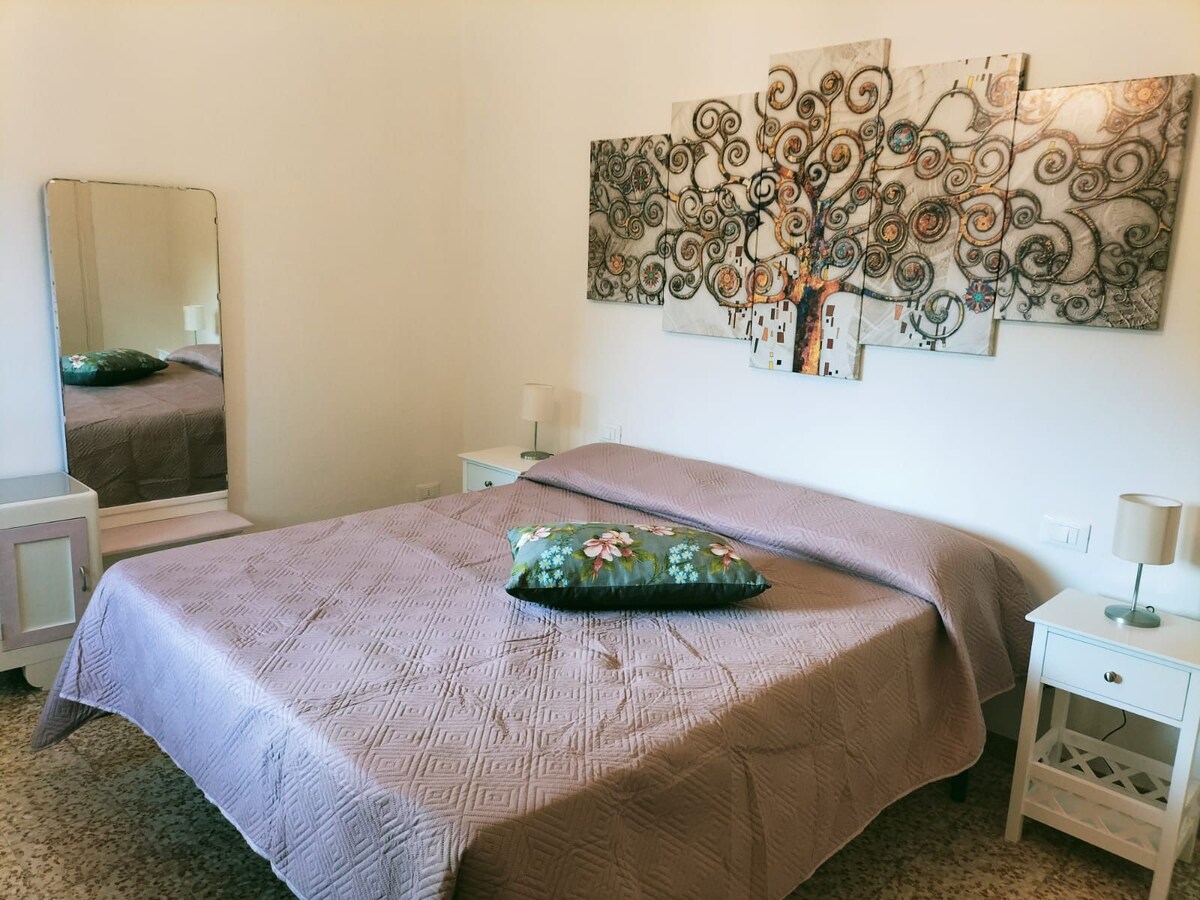 The sleeping area features a spacious bed covered with a patterned blanket and a decorative pillow. A mirrored wardrobe reflects soft light, while bedside tables with lamps flank the bed. A colorful wall art piece depicting an abstract tree adds visual interest to the room.