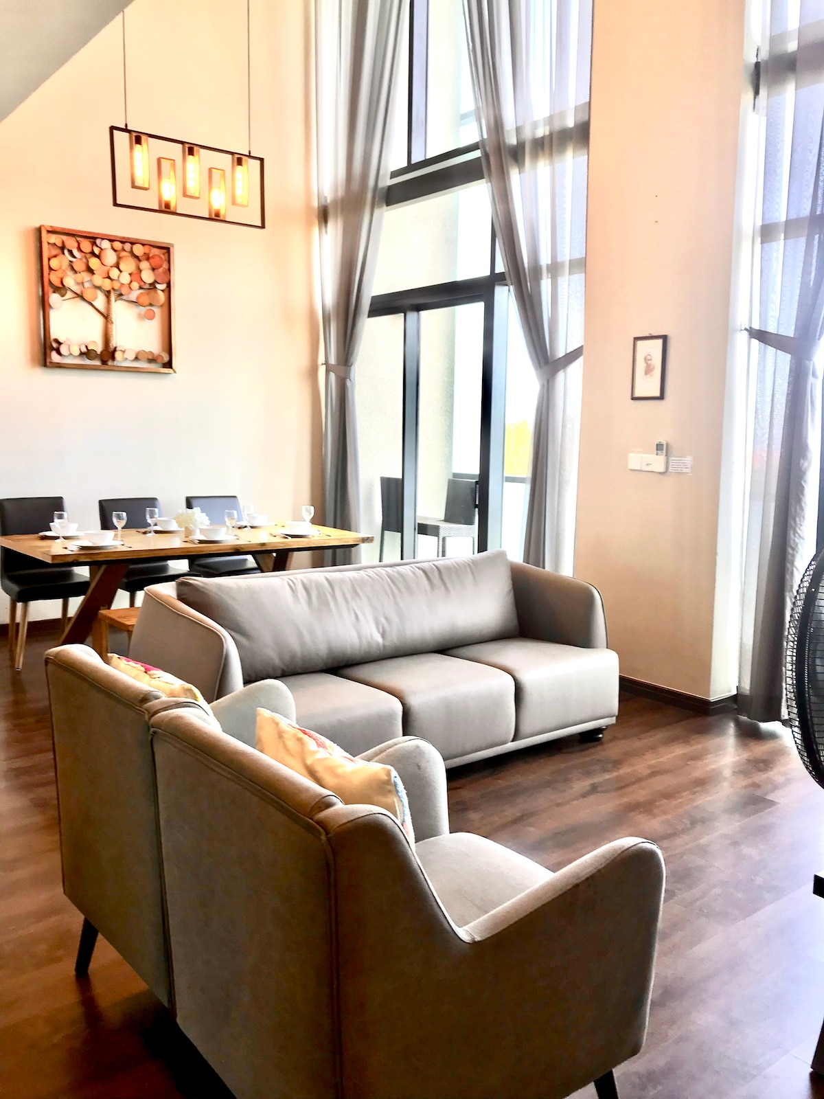 A spacious living area features large windows allowing natural light to fill the room. Comfortable grey sofas are arranged invitingly. A dining table set for six is visible in the background, with elegant artwork adorning the wall, enhancing the modern decor.