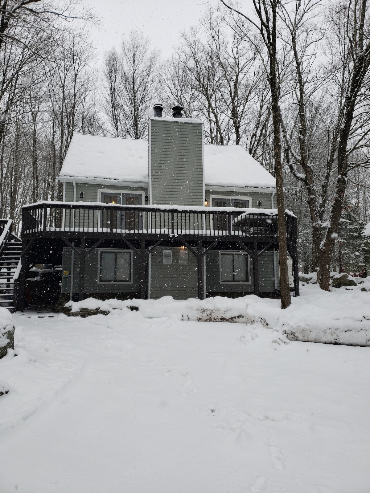 A charming cabin stands amidst a snowy landscape, showcasing a two-story structure with a wrap-around deck. Snow blankets the ground and roof, while trees in the background create a serene winter scene. Large windows invite natural light into the inviting space.