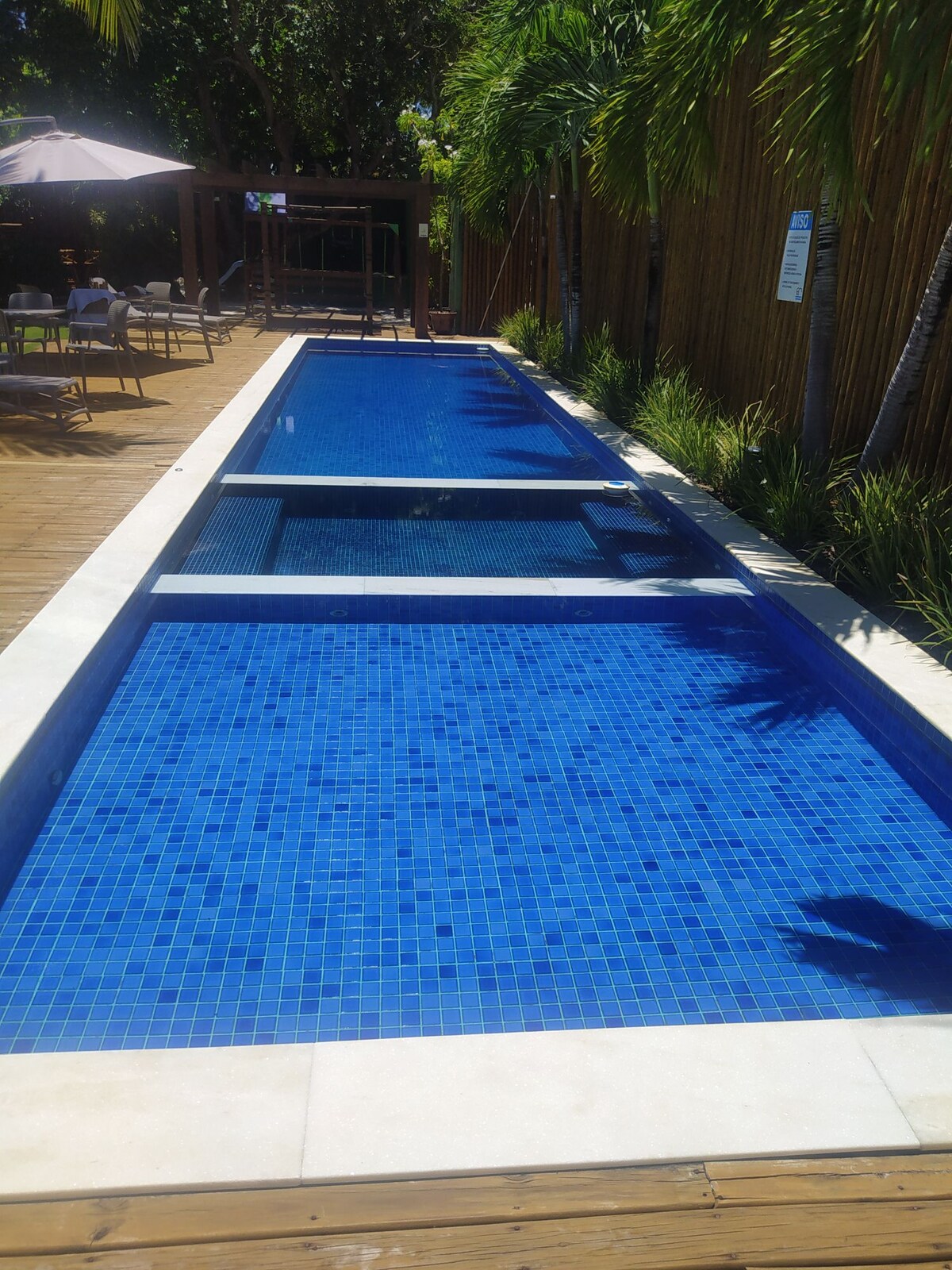 An outdoor pool featuring clear blue water is surrounded by lush greenery and wooden decking. The pool includes distinct sections for recreation, with spacious areas for lounging visible in the background. Shade is provided by nearby umbrellas, contributing to a serene atmosphere.