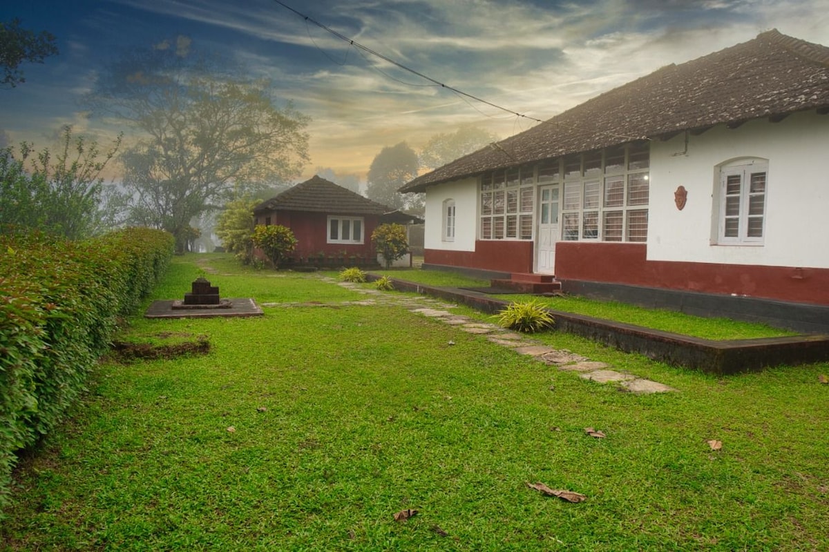 A serene garden area is framed by well-maintained grass and lush greenery. Two rustic buildings are visible, featuring large windows and traditional architecture. Soft morning light filters through the trees, creating a calm and inviting outdoor space.