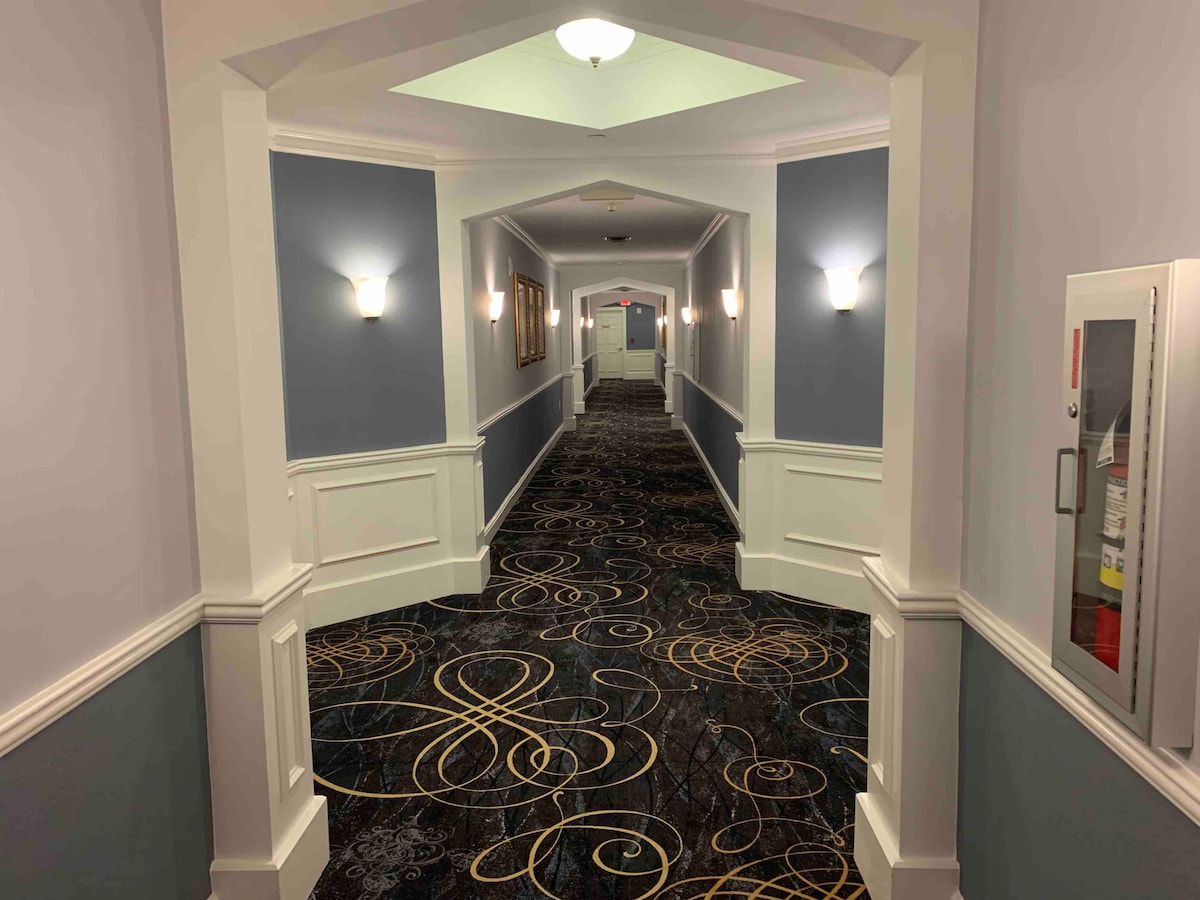 A spacious hallway is illuminated by wall sconces along light blue walls. A patterned carpet with swirling designs leads to a doorway at the end, framed by white railings. The soft lighting creates a welcoming and peaceful atmosphere.