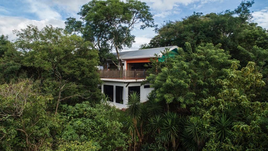 A view of the house is captured from above, surrounded by lush greenery. The structure features a spacious terrace with a railing, visible in the foreground. Dense trees and various plants create a natural backdrop, emphasizing the property's seclusion and connection to nature.