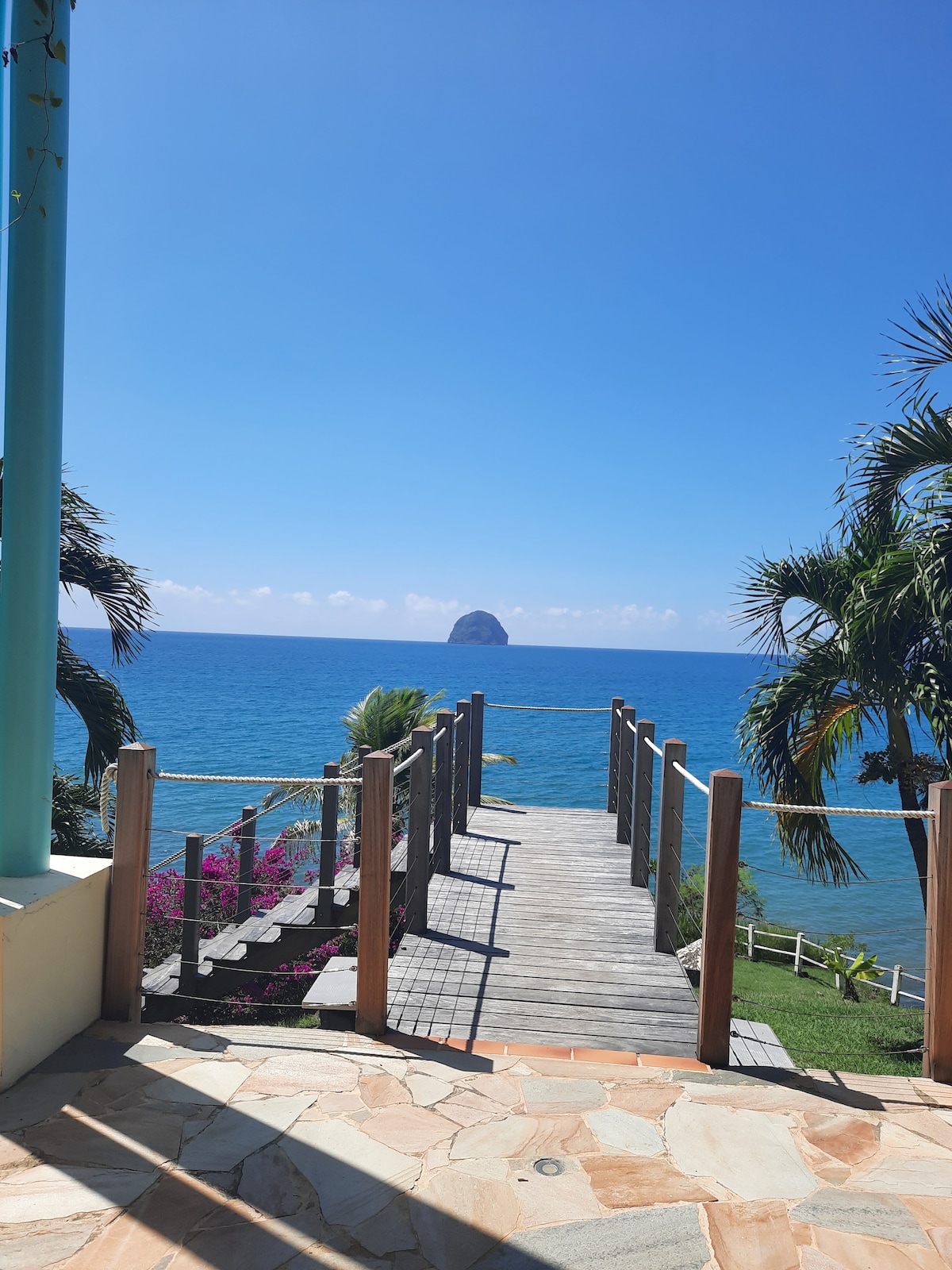 A wooden pathway leads toward the turquoise sea, framed by lush greenery and blooming flowers. The iconic rock of Diamant is visible in the distance, rising gently from the ocean under a clear blue sky.