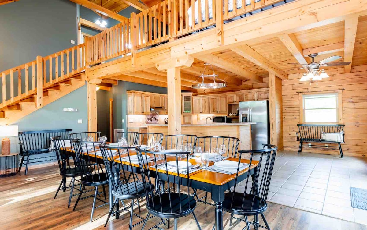 A spacious dining area features a long wooden table surrounded by black spindle-back chairs. An open kitchen is visible in the background, equipped with modern appliances and wooden cabinetry. Natural light streams in through windows, illuminating the wood-paneled ceiling and inviting warmth into the space.