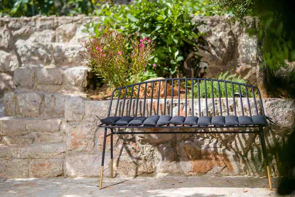 A modern black bench with cushioned seating is positioned against a rustic stone wall. Lush greenery and flowering plants surround the area, creating a serene outdoor space. Sunlight casts gentle shadows on the bench, enhancing the tranquil atmosphere.