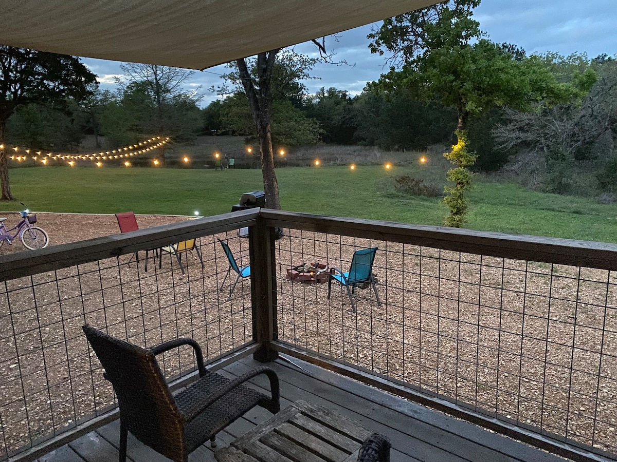 A view from a covered deck shows a spacious outdoor area with string lights illuminating a gathering space. Colorful chairs surround a fire pit, and an open grassy field extends beyond, suggesting a serene setting for relaxation.