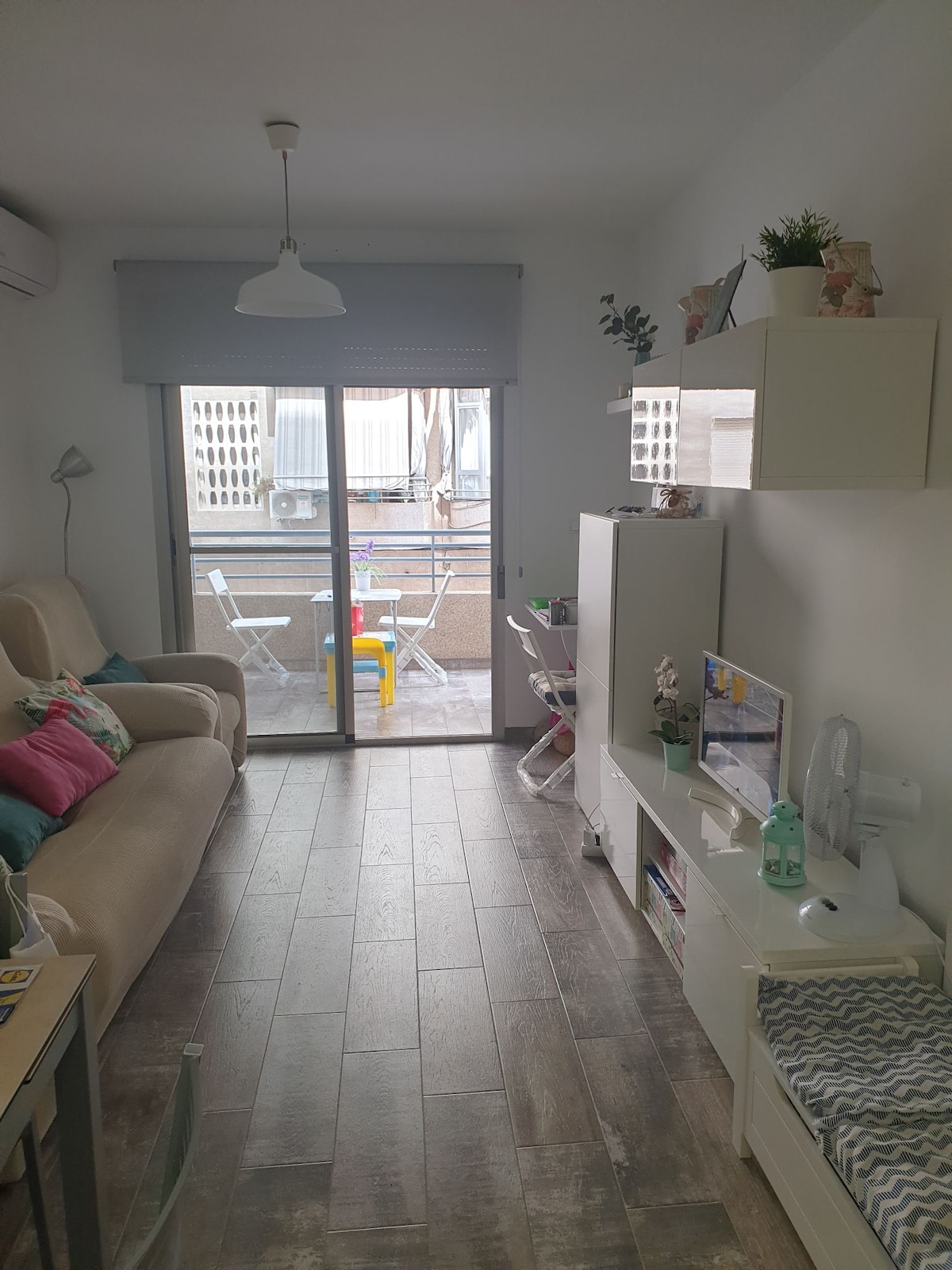 A bright living area features a light-coloured sofa with assorted cushions. A sliding glass door leads to an outdoor space, while shelves display decorative items. A compact television and desk are positioned along the wall, offering functionality and comfort.