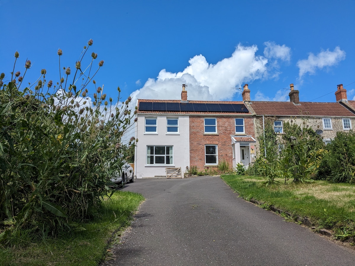 A charming family home is depicted with a blend of traditional and modern design. Solar panels adorn the roof, while a spacious driveway leads to the entrance. Lush greenery frames the scene, highlighting the inviting surroundings under a bright blue sky dotted with clouds.