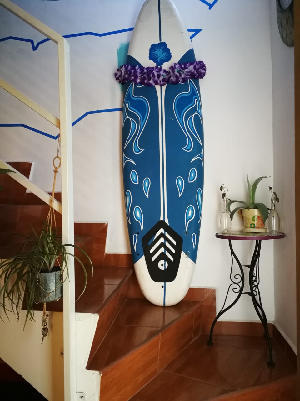 A blue surfboard adorned with a floral lei is positioned against the wall near a staircase. The setting features warm-toned tiles and a small table with a plant and glass drinkware, contributing to a relaxed coastal vibe.