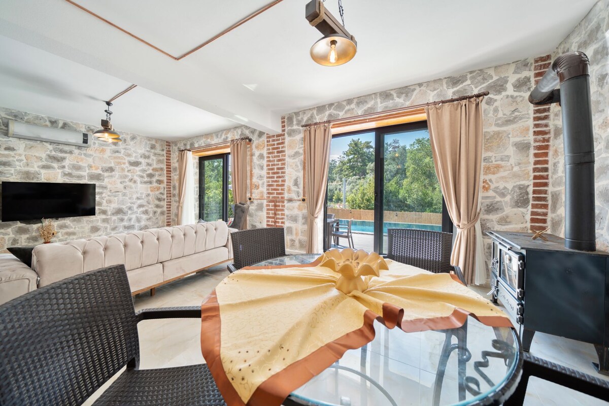 An open living space features a circular glass dining table with a decorative tablecloth. A cozy seating area includes a plush couch and is complemented by a television mounted on a stone wall. Large windows offer views of the surrounding nature, with curtains framing the openings.