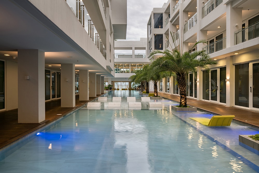 A tranquil swimming pool is framed by modern architecture, with lounge chairs positioned beside the water. Palm trees provide greenery along the poolside, while soft lighting creates a soothing atmosphere. The pool is designed with a sleek surface, reflecting the surrounding buildings and sky.