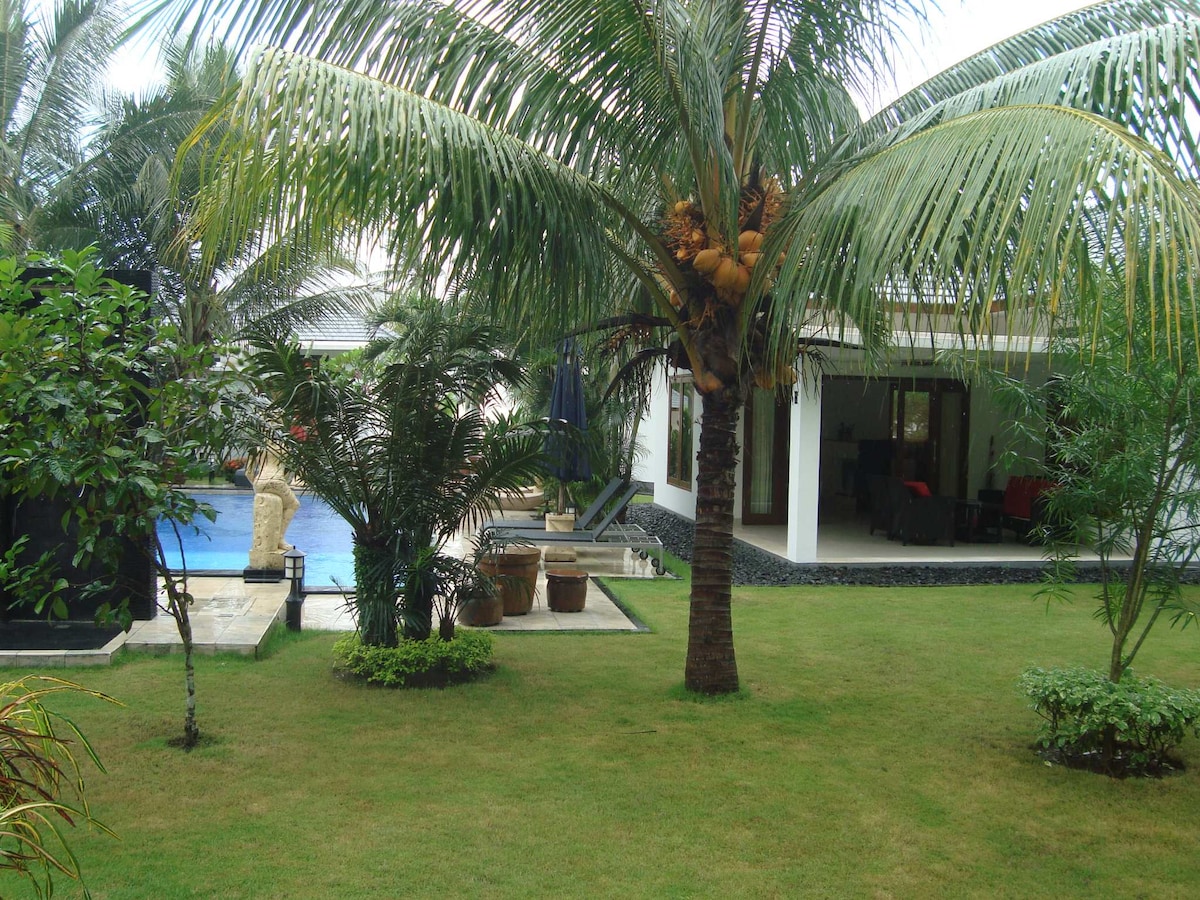 A serene garden view is presented, featuring lush greenery, palm trees, and flowering plants. The outdoor pool is visible in the distance, surrounded by a stone terrace. A covered seating area is included, providing a relaxing spot adjacent to the vibrant landscape.