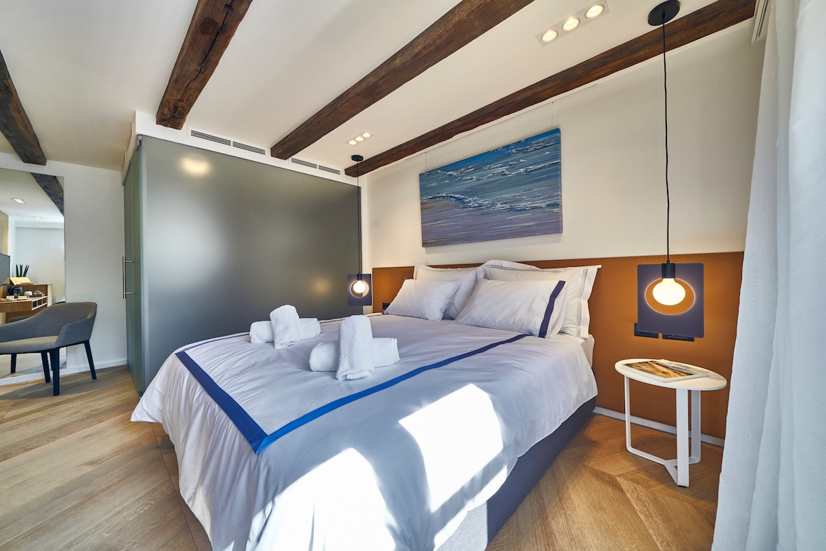 A spacious bedroom features a double bed adorned with soft linens and neatly arranged towels. Modern lighting hangs above the bed, illuminating the artwork on the wall. Natural light filters through a nearby window, highlighting the room's warm wooden beams and contemporary decor.