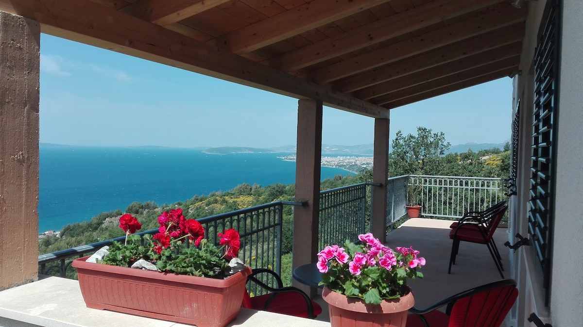 A covered terrace offers a view of the sea and distant city. Decorative flower boxes filled with vibrant blooms frame the space. Two red chairs are positioned beside a sturdy table, inviting relaxation. The natural setting is complemented by a wooden ceiling and metal railing.