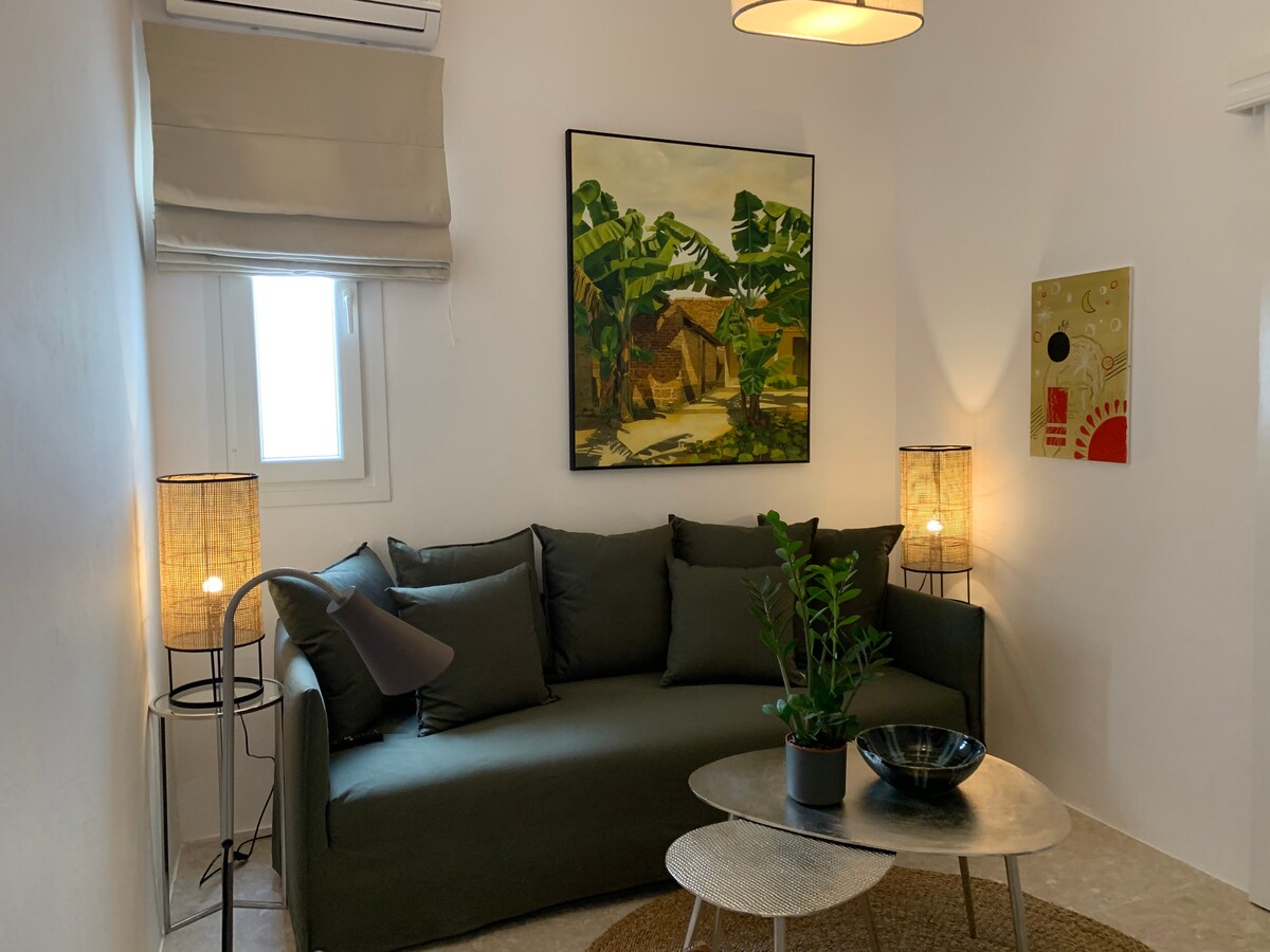 A cozy seating area is furnished with a green sofa adorned with dark cushions. A round coffee table sits centrally, accompanied by decorative items. Soft lighting is provided by floor lamps, and artwork featuring vibrant colors adorns the walls.