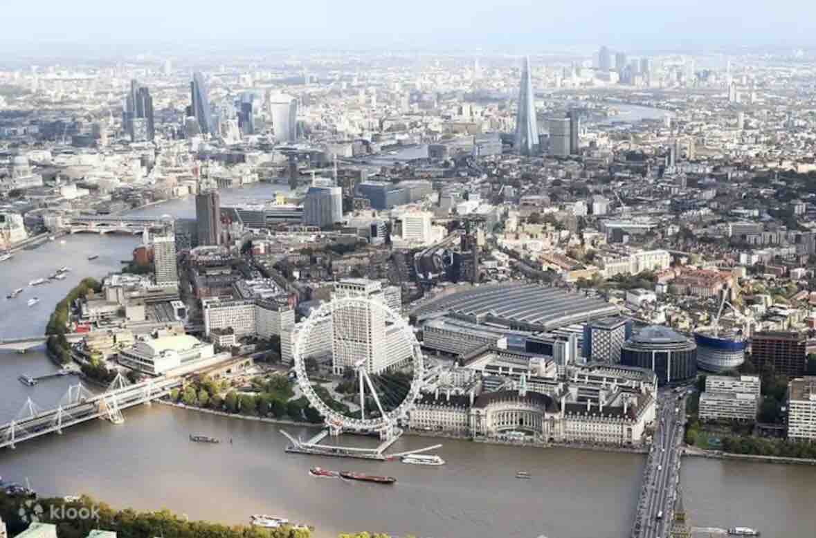 An aerial view captures the sprawling cityscape of London, with the River Thames flowing through it. Iconic landmarks, including the London Eye and modern skyscrapers, showcase the blend of history and contemporary architecture. Boats dot the water, and the landscape is framed by distant rolling hills.