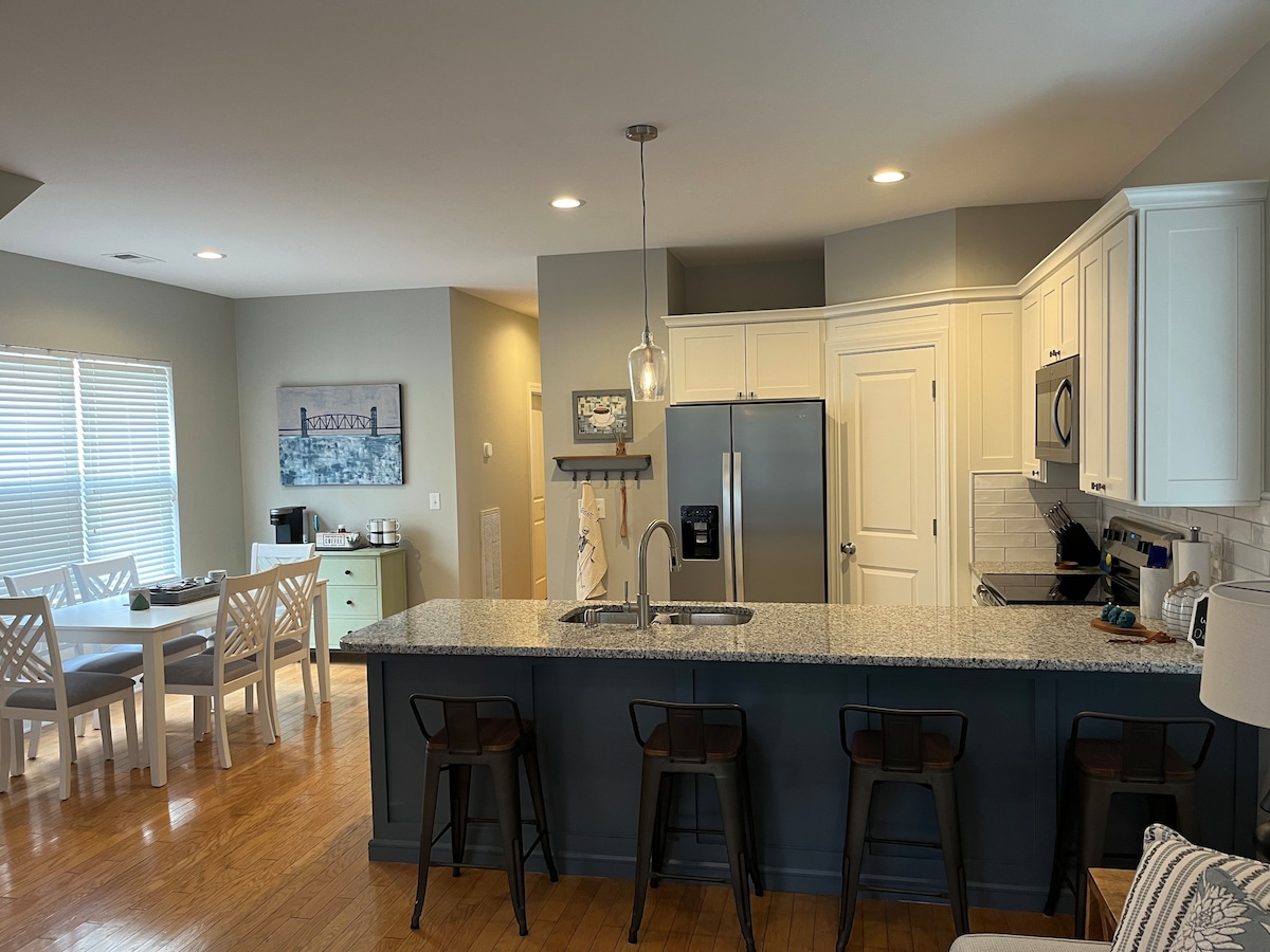 A modern kitchen features granite countertops and white cabinets. A stainless steel refrigerator and microwave are visible. Bar seating for four is provided at the countertop. Natural light fills the space from a nearby window, with a dining area accommodating six visible in the background.