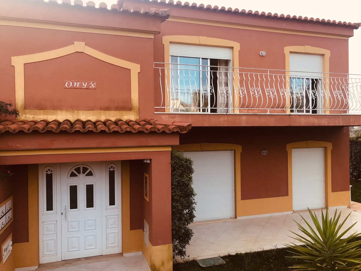 The exterior of the two-story apartment building showcases a warm terracotta facade with decorative trim. A covered entrance leads to the main door. Large windows with shutters are visible above, and a balcony with wrought iron railings complements the entrance. Lush greenery surrounds the property.