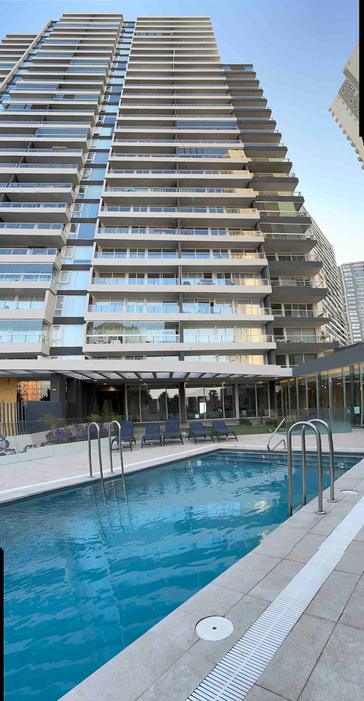 A modern building stands tall with multiple floors, showcasing expansive glass balconies. The outdoor pool is surrounded by stylish lounge chairs, and the nearby indoor pool is visible through large windows, inviting relaxation.