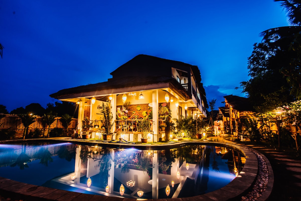 The exterior of the homestay is illuminated at night, showcasing a cozy outdoor swimming pool and a spacious terrace adorned with lush greenery. The building's architecture blends traditional and modern elements, inviting guests to enjoy the evening ambiance surrounded by nature.