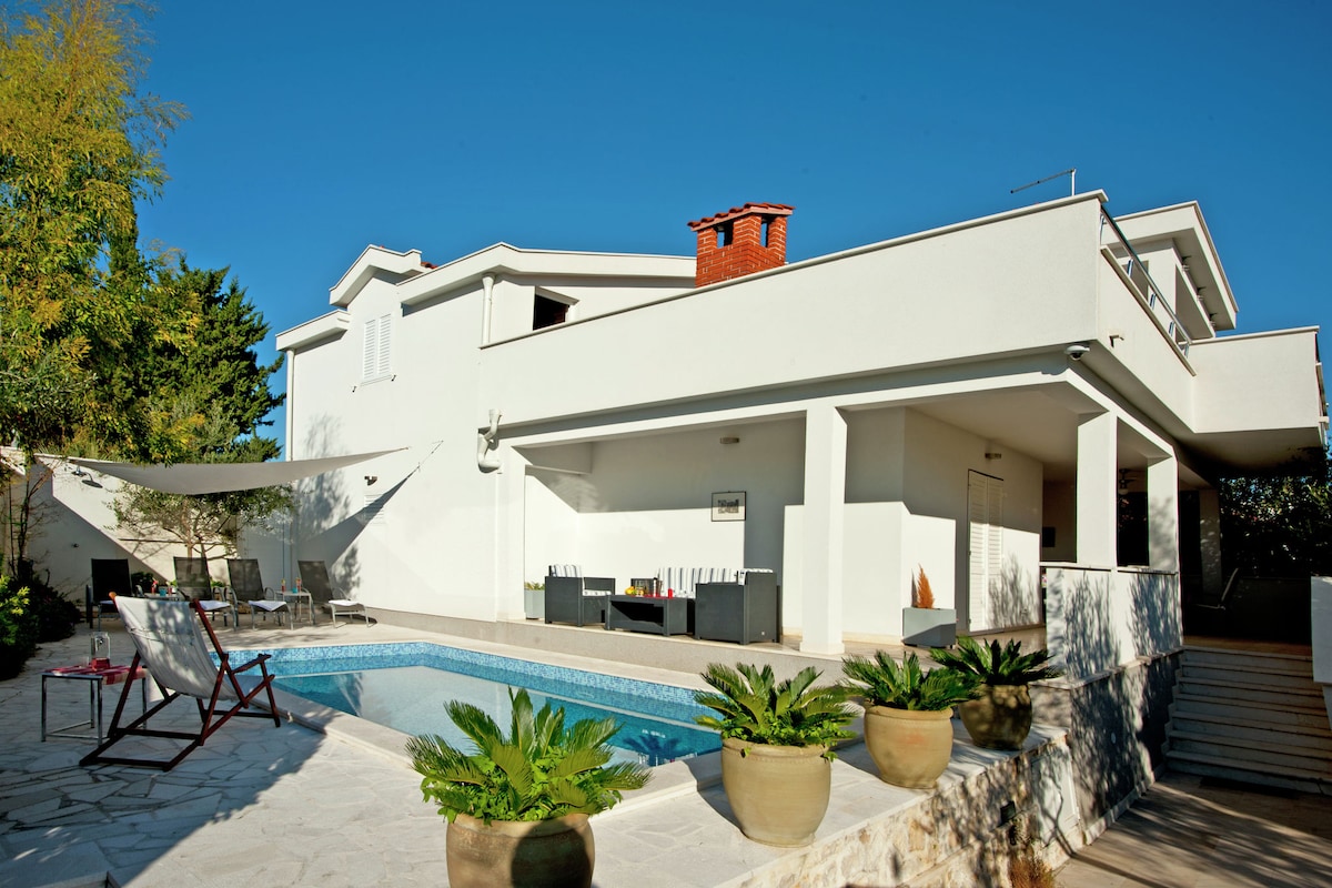 The exterior of the property features a modern two-story house with a terrace and a private swimming pool. Potted plants are arranged around the pool area, and seating options are present under a shaded area. Clear blue skies set a bright backdrop for the scene.