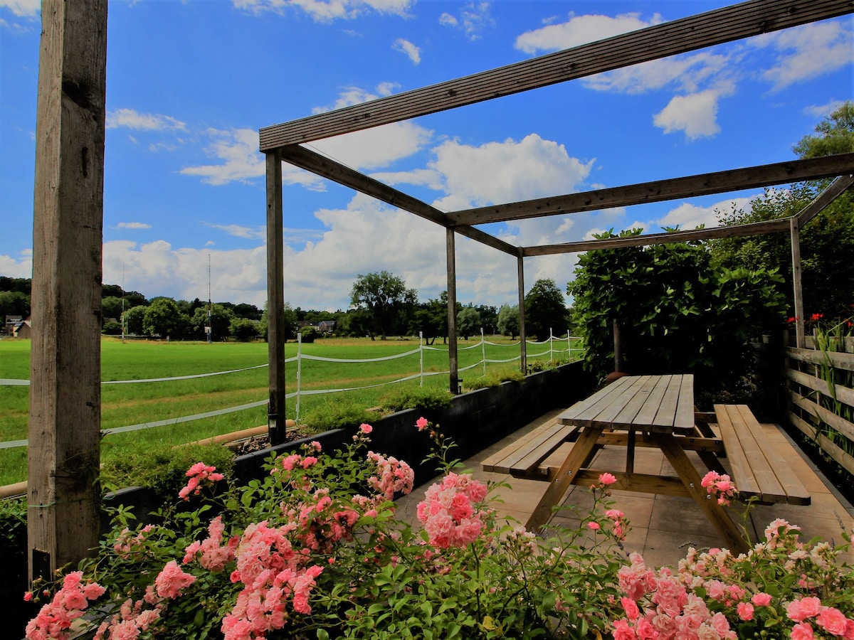 An outdoor seating area is surrounded by vibrant pink flowers. A wooden table with attached benches is positioned beneath a pergola, providing shade. The view features lush green fields and a clear blue sky with scattered clouds in the background.