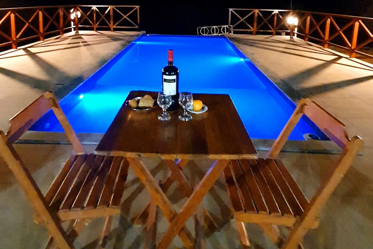 A small wooden table is set with a bottle of wine, glasses, bread, and a piece of fruit, positioned next to a blue-lit pool. Two folding chairs are arranged around the table, with wooden railings visible in the background, enhancing the inviting outdoor space.