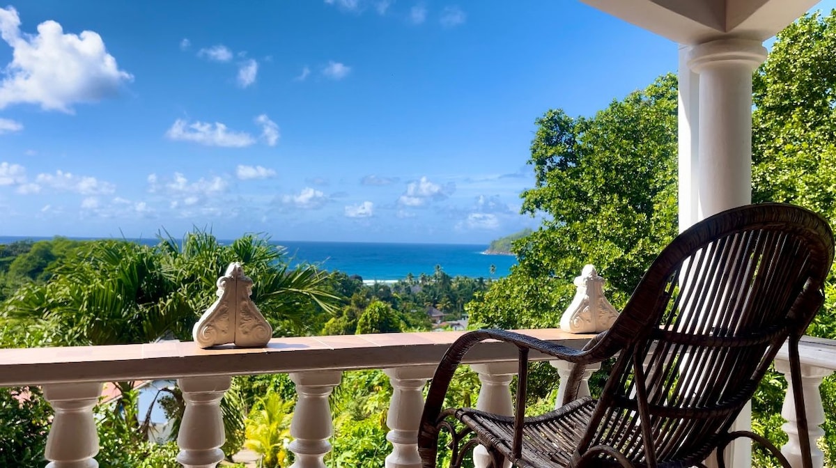 A private balcony features a cozy wicker chair, positioned to offer a clear view of the ocean and surrounding greenery. Lush trees frame the scene, while the sky is dotted with fluffy clouds, enhancing the tranquil seaside atmosphere.
