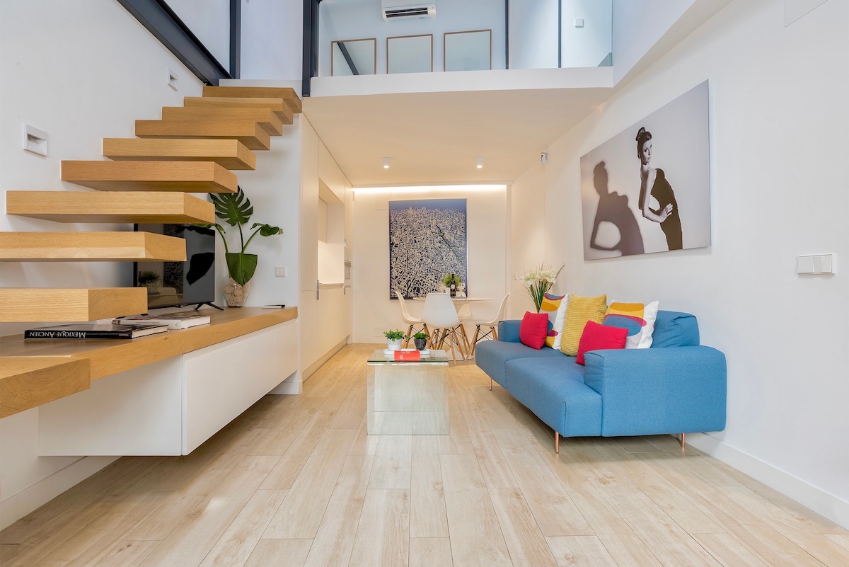 A modern living space features a light wooden staircase leading to an upper level. A white wall showcases a black-and-white artwork, while a cozy blue sofa with colorful throw pillows is positioned beside a glass coffee table. Abundant natural light illuminates the area.