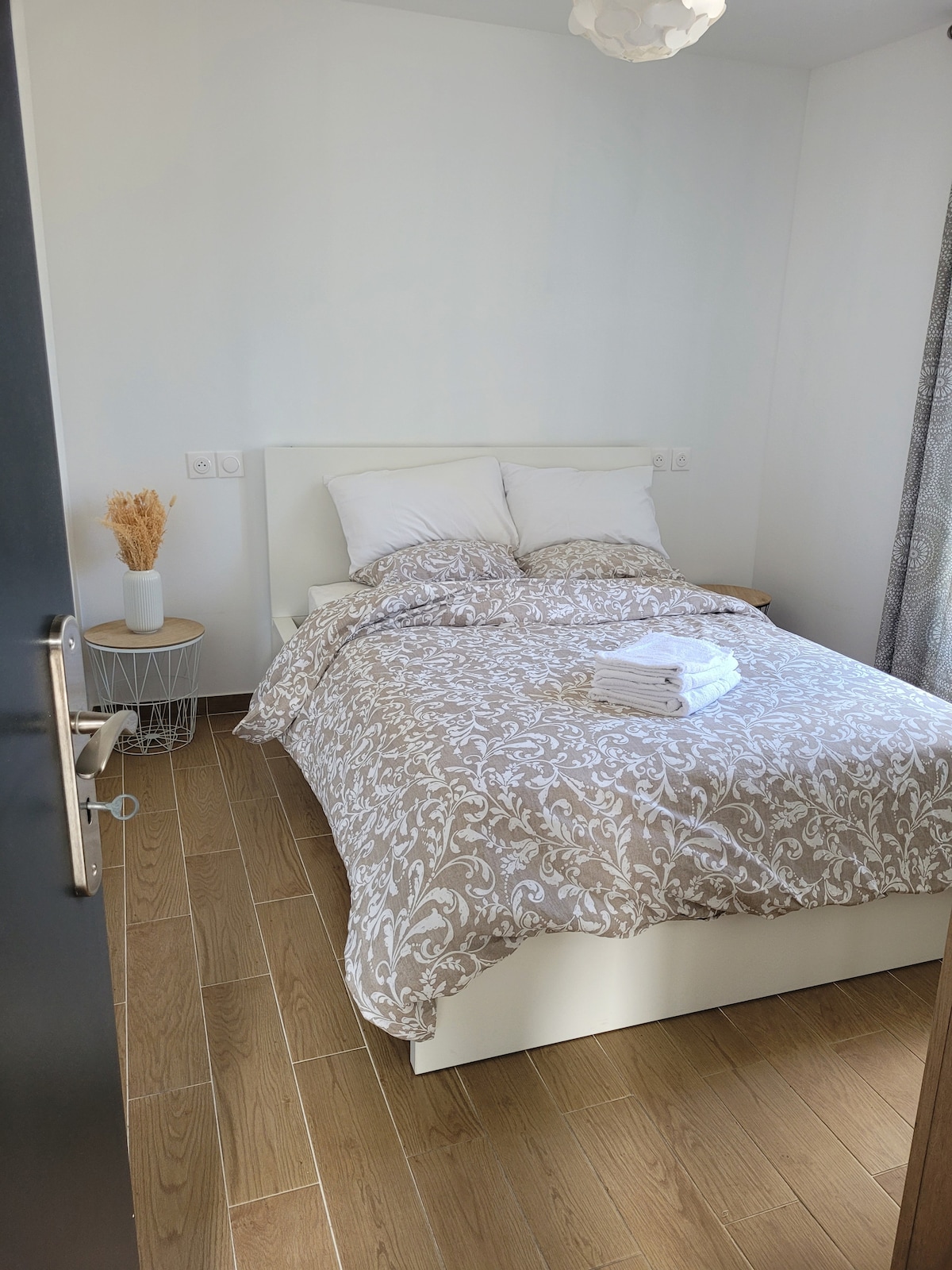 A tranquil bedroom is furnished with a spacious bed topped with a patterned duvet and neatly arranged pillows. Fresh towels are placed on the bed, and a small side table with a decorative vase adds an elegant touch. Natural light filters through the window, enhancing the serene ambiance.