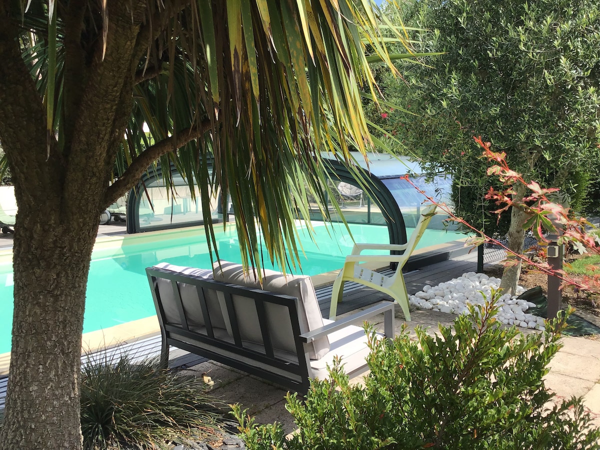 A serene outdoor space is presented, featuring a swimming pool partially covered, surrounded by lush greenery and palm trees. Comfortable seating, including a white couch and an armchair, is situated nearby, inviting relaxation in this shaded area.