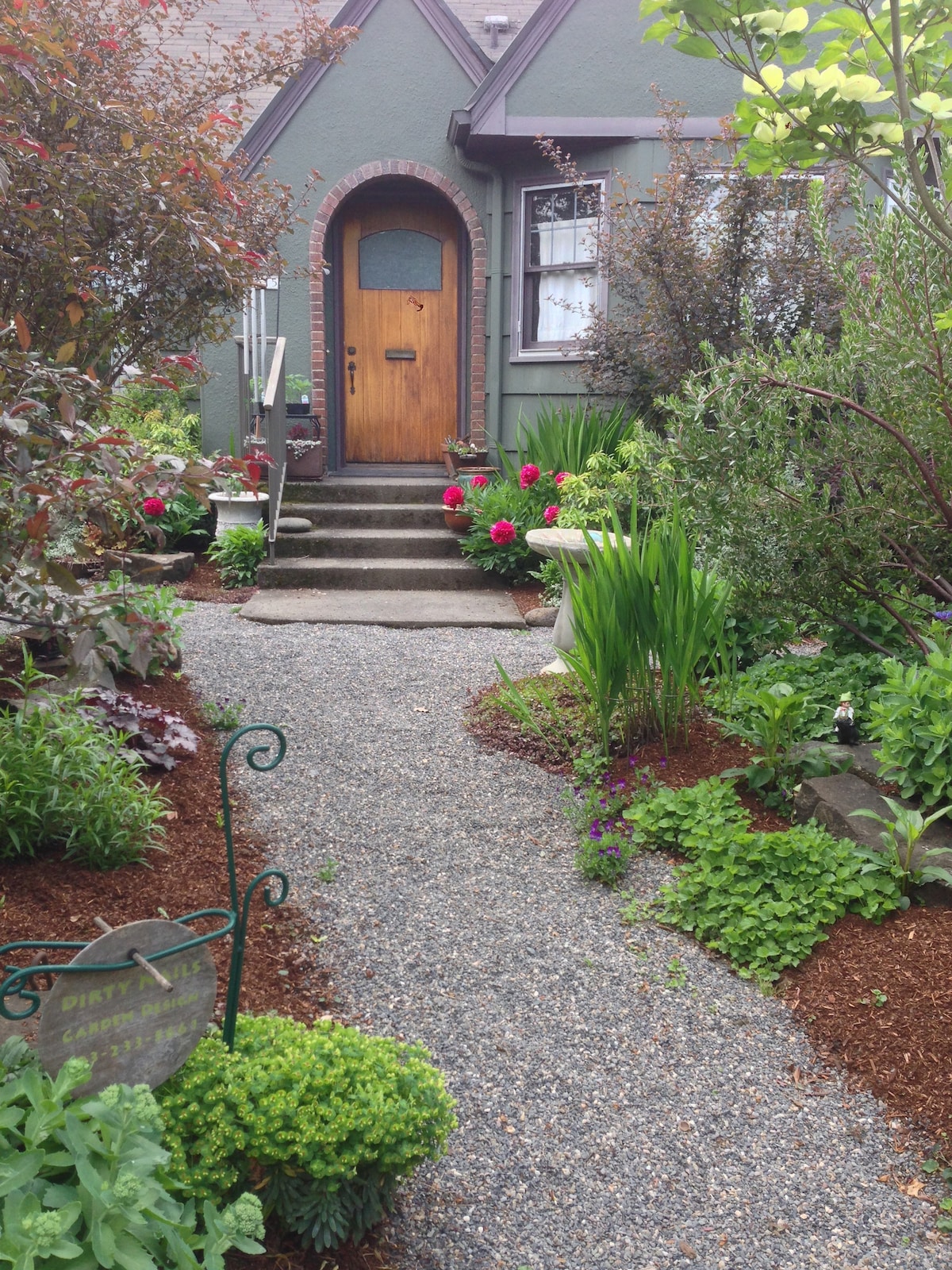 A garden path made of gravel leads to a wooden front door framed by a variety of colorful flowers and lush greenery. The surrounding garden features well-maintained plants, including bushes and decorative rocks, creating a welcoming entrance to the studio.