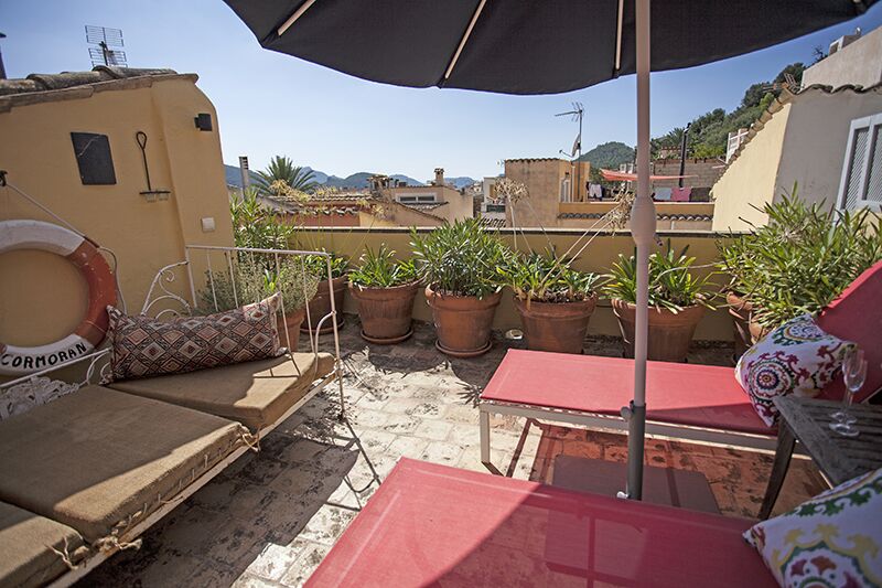 A sunlit terrace features comfortable lounge chairs and a small table surrounded by potted greenery. An umbrella provides shade, while rooftops and distant mountains create a scenic backdrop. Colorful cushions add a relaxed touch to the space.