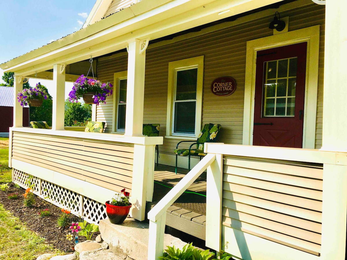 The exterior of Corner Cottage features a welcoming porch with hanging flower baskets and a decorative sign. Two chairs are positioned on the porch, complemented by vibrant plants in containers, creating an inviting entryway.