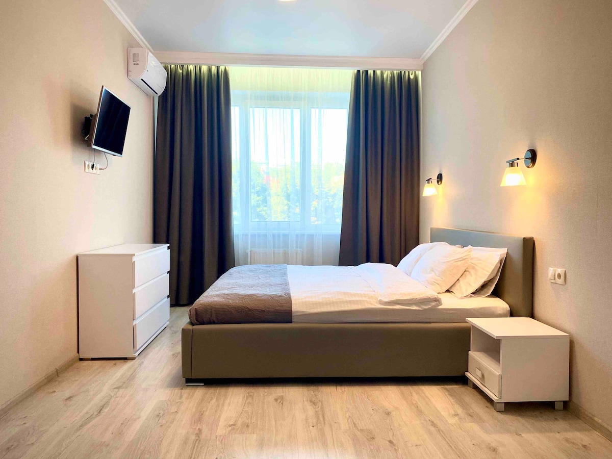 A spacious sleeping area is depicted, featuring a comfortable bed with light bedding. A dresser and nightstand are present. Large windows with sheer curtains allow natural light to fill the room, complemented by soft wall sconce lighting.