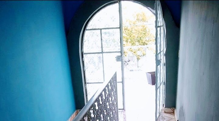 A blue-painted corridor is illuminated by natural light filtering through a large arched door with glass panels. A patterned railing provides a view of the outdoor space just beyond the door.