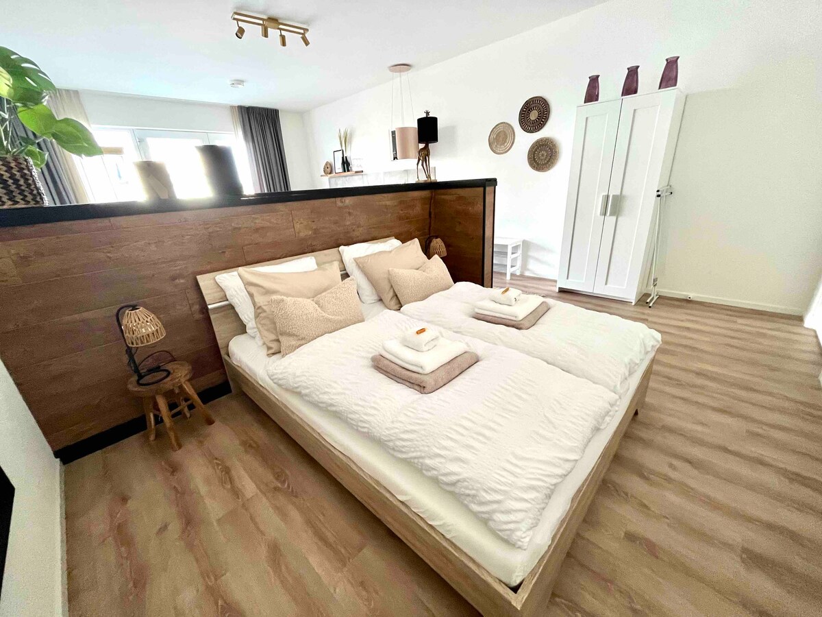 A spacious sleeping area features a comfortable bed with soft bedding and arranged towels. The room is complemented by wooden accents, natural light flooding in through large windows, and minimalistic decor, enhancing a relaxing environment.