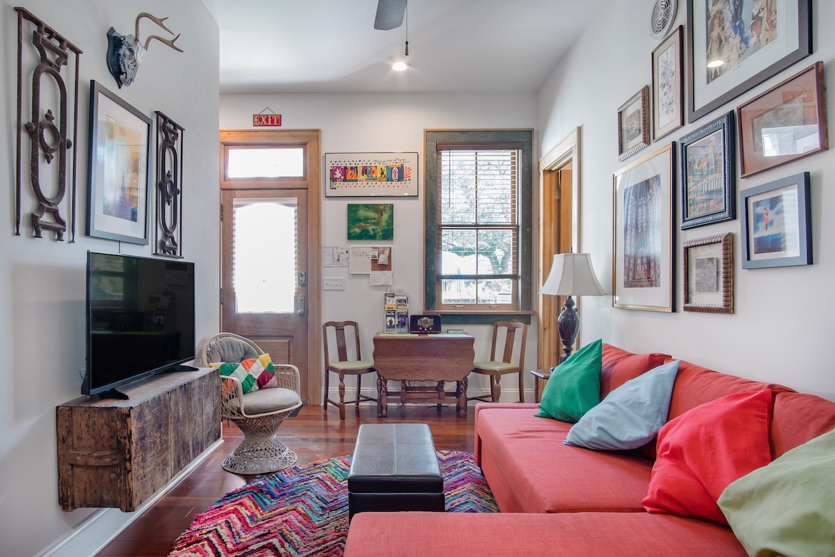 A vibrant sitting area with a coral sofa adorned with colorful cushions is featured. A rustic wooden television stand holds a TV, and eclectic artwork decorates the walls. At the back, a small dining table sits next to a door that provides natural light.